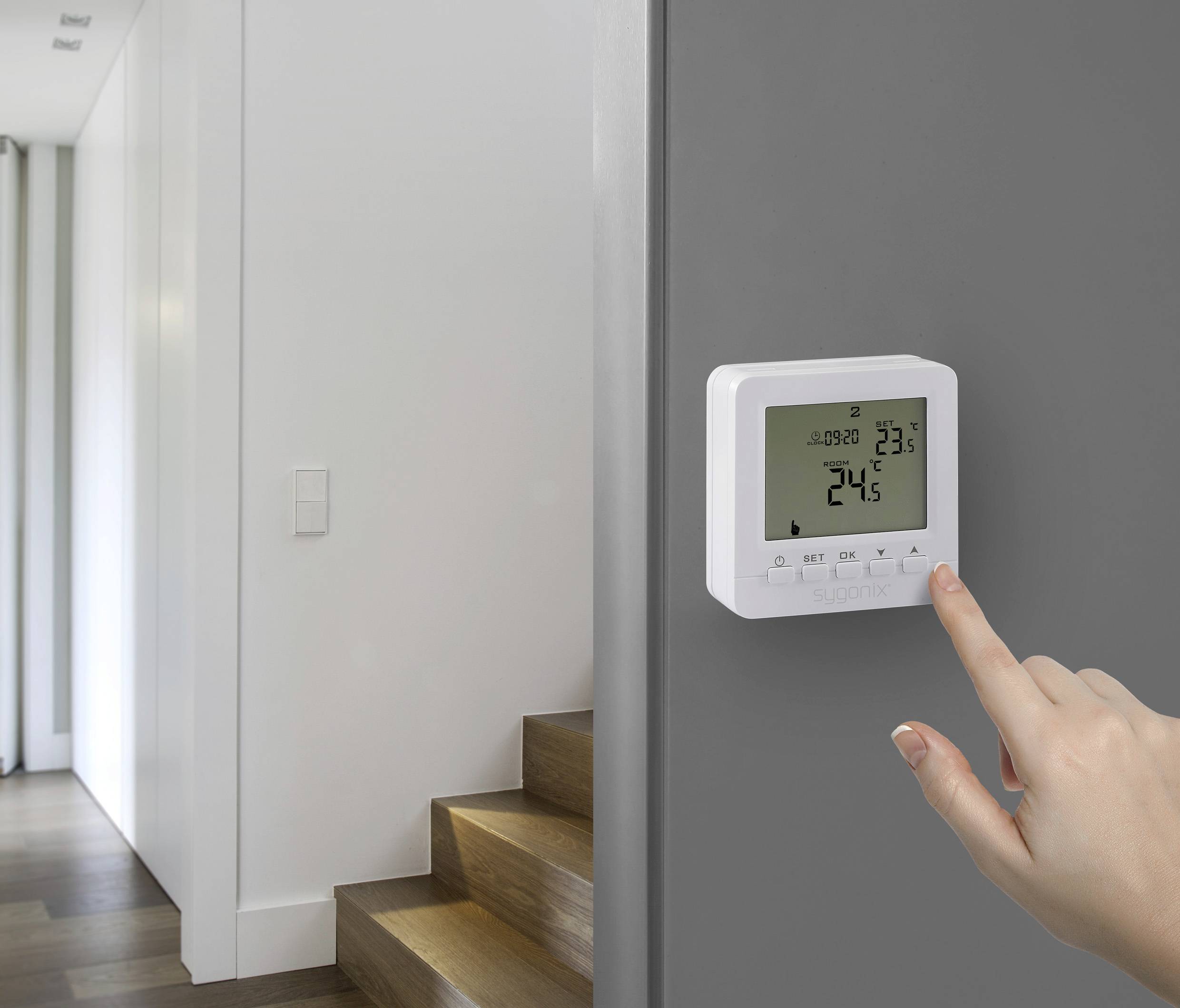 Un main ajuste un thermostat numérique affichant '24,5°C'. Il est fixé sur un mur gris à côté d'un escalier en bois dans une maison moderne.