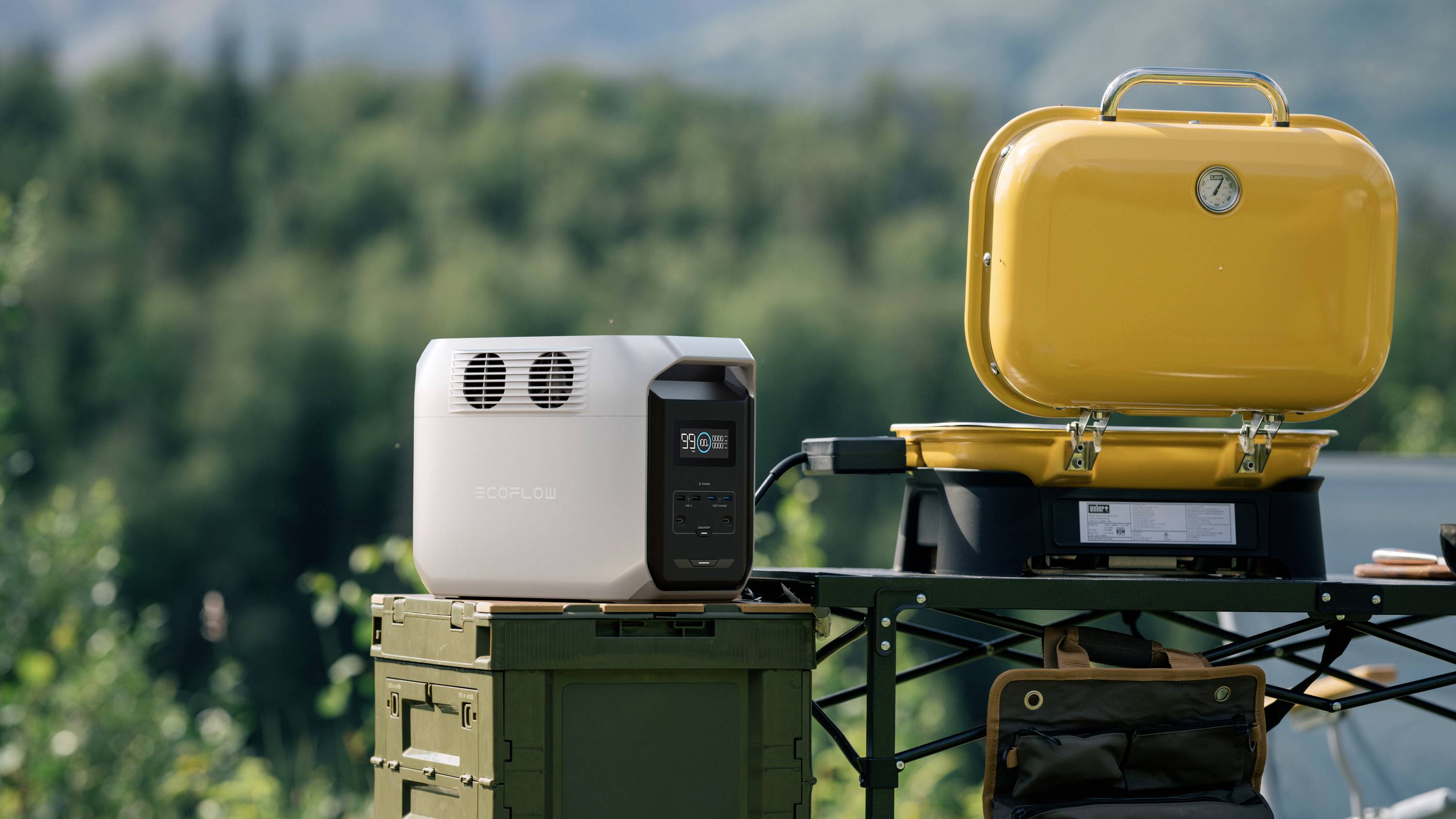 Un générateur portable est posé sur une table à l'extérieur, à côté d'un grill jaune. En arrière-plan, on aperçoit un paysage boisé.