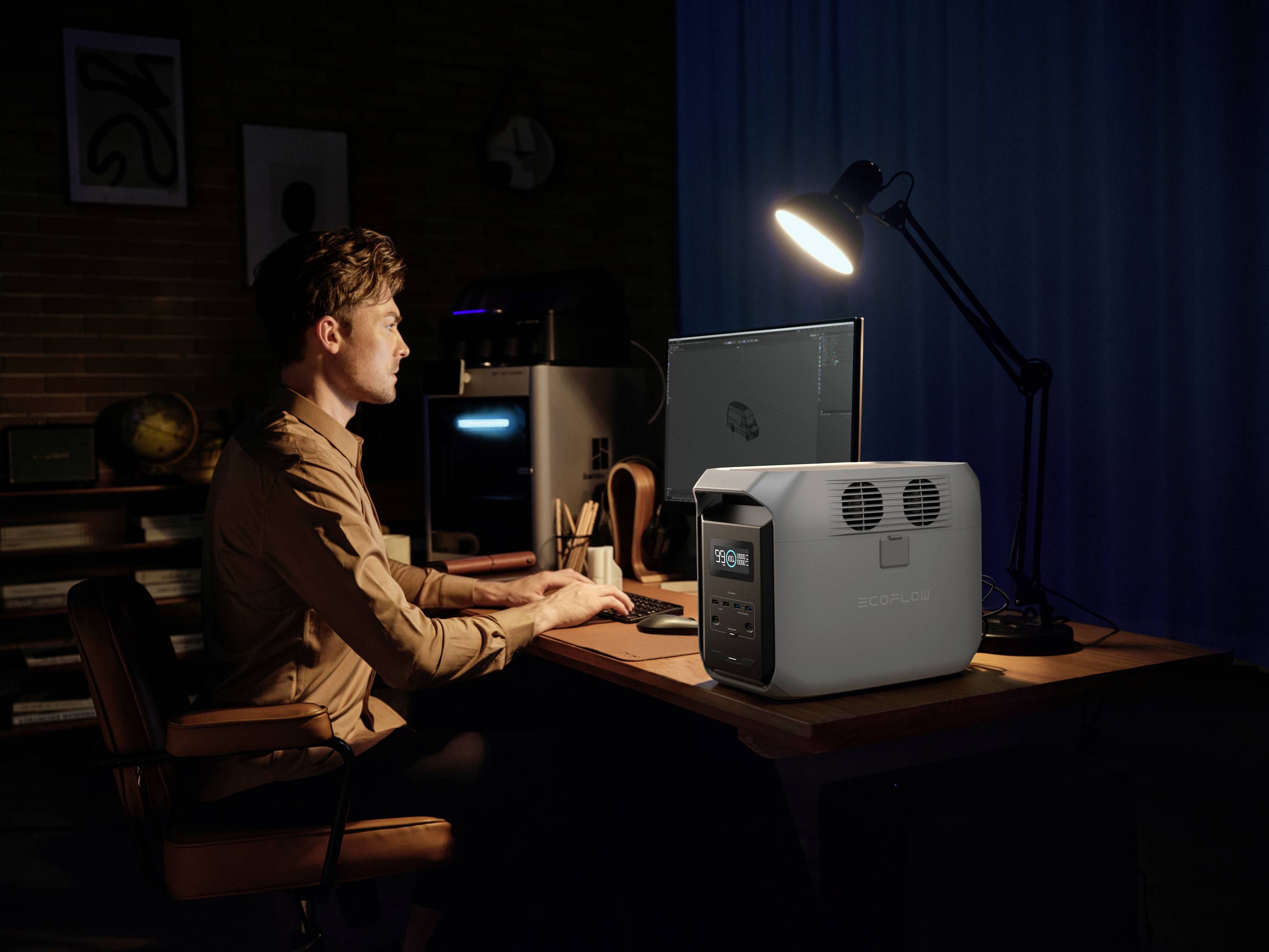 Une personne travaille le soir sur un ordinateur dans un bureau. Un grand accumulateur avec un affichage numérique est posé sur le bureau. La pièce est faiblement éclairée.