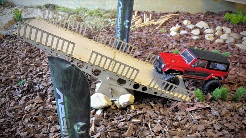 Une petite voiture de modélisme rouge roule sur une rampe inclinée dans un paysage miniature composé de copeaux de bois bruns et de petits rochers.