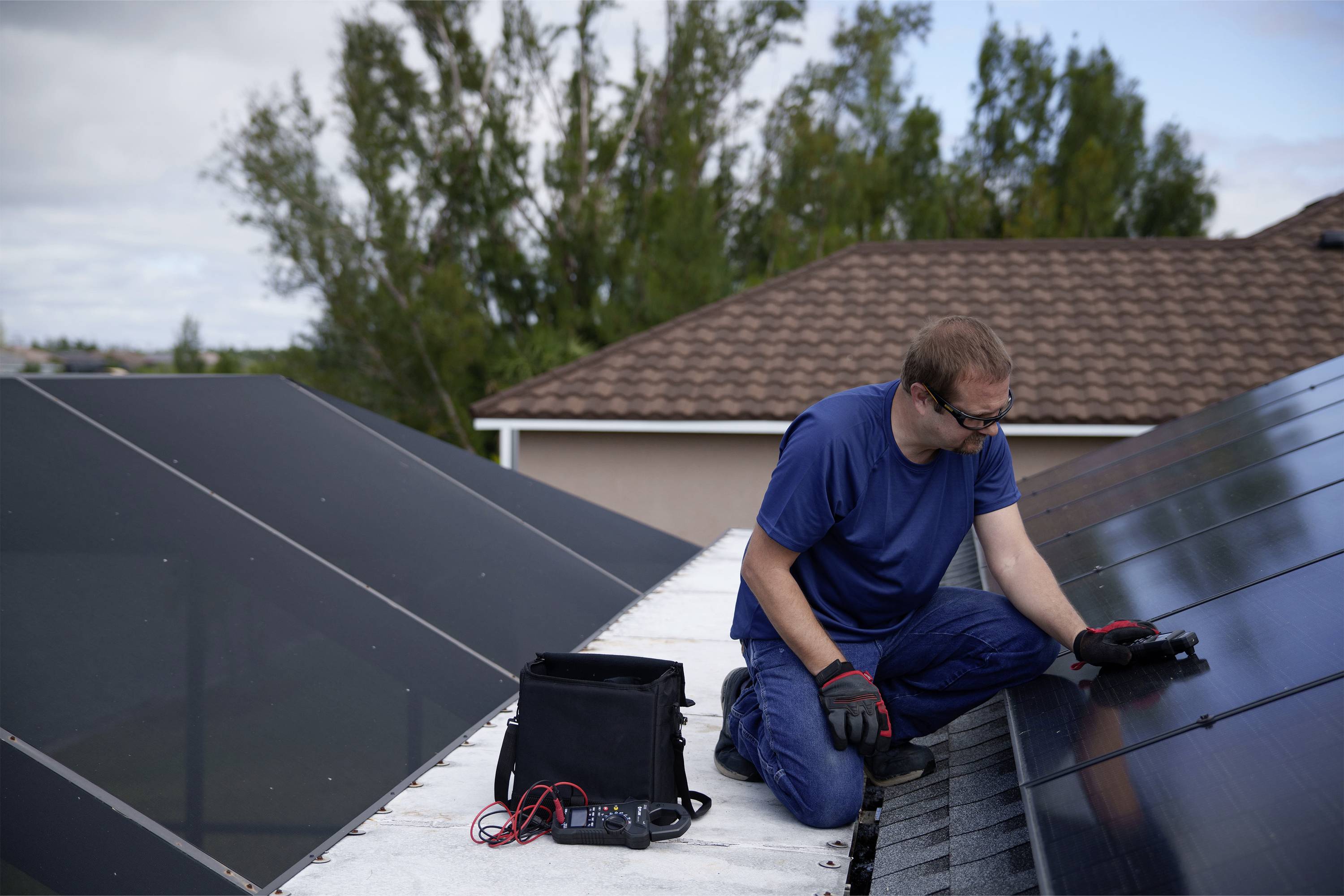Un homme en vêtements bleus travaille sur des modules solaires sur un toit, avec des outils et un sac à proximité ; des arbres et le ciel en arrière-plan.