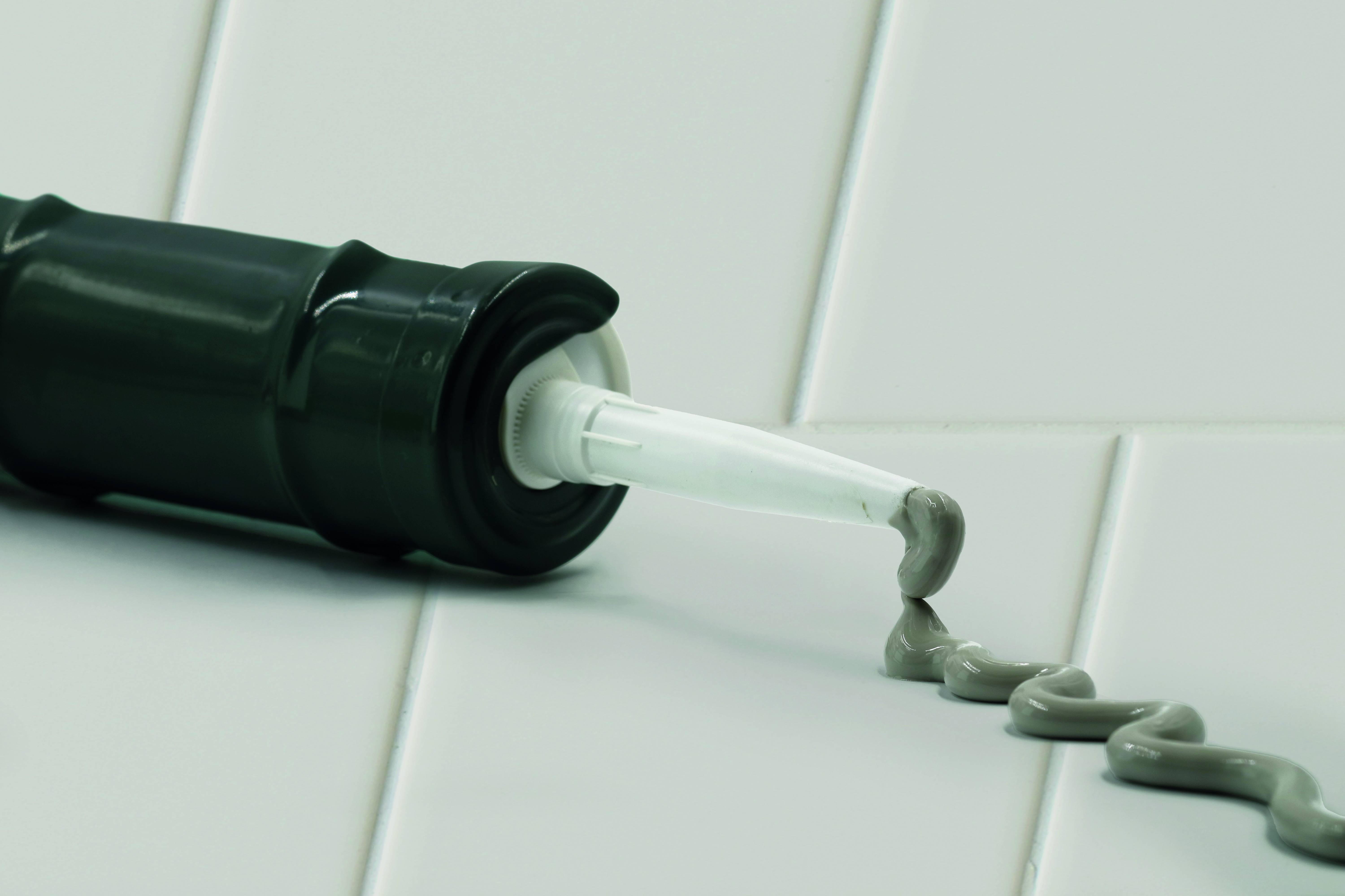 Gros plan d'un tube noir distribuant un mastic gris sur une surface carrelée blanche, montrant un cordon d'adhésif en cours d'application.