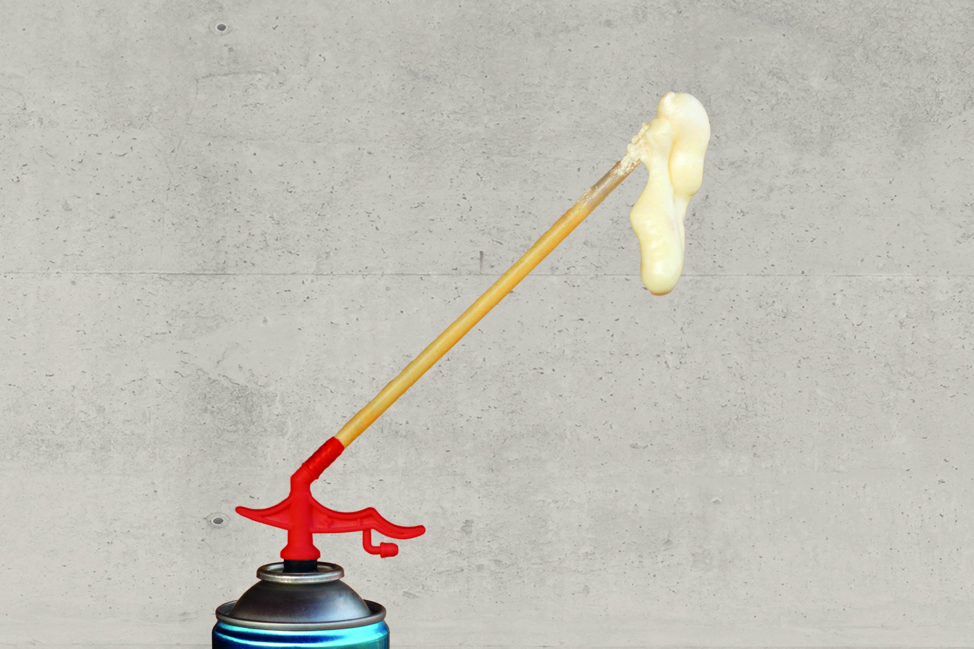 Un spray attaché à une longue paille projette de la mousse vers le haut et vers la droite contre un mur gris uni.