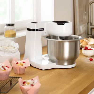 Robot de cuisine sur une table en bois, entouré d'ingrédients tels que de la farine, des œufs et des baies. Des muffins prêts sont disposés dans des moules roses.