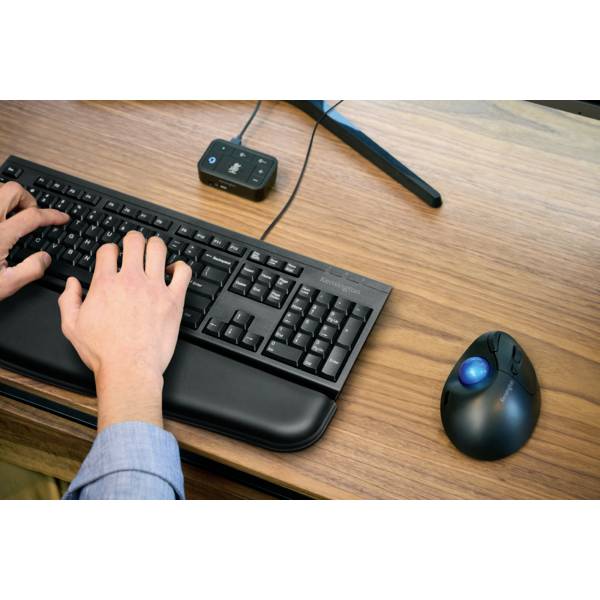 Deux mains tapent sur un clavier d'ordinateur noir posé sur une table en bois, à côté desquelles se trouvent une souris ergonomique et un microphone.