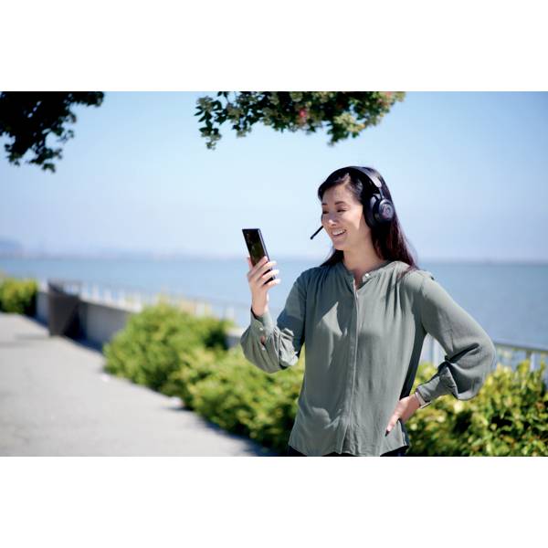 Une personne se tient dehors près d'un plan d'eau, porte des écouteurs avec microphone et sourit en regardant son smartphone.