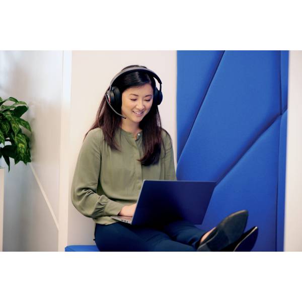 Femme souriante portant des écouteurs, travaillant sur un ordinateur portable, assise sur un banc bleu avec une plante en arrière-plan.