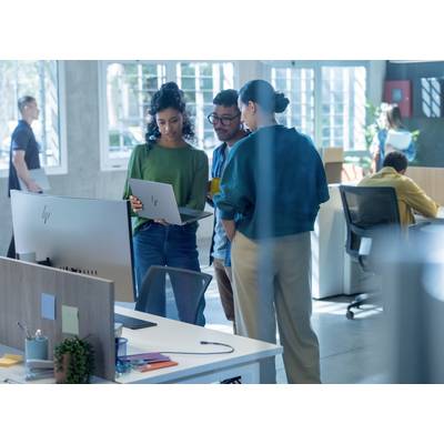 Trois personnes sont debout dans un bureau moderne et regardent ensemble un ordinateur portable. À l'arrière-plan, on peut voir des bureaux et des fenêtres.