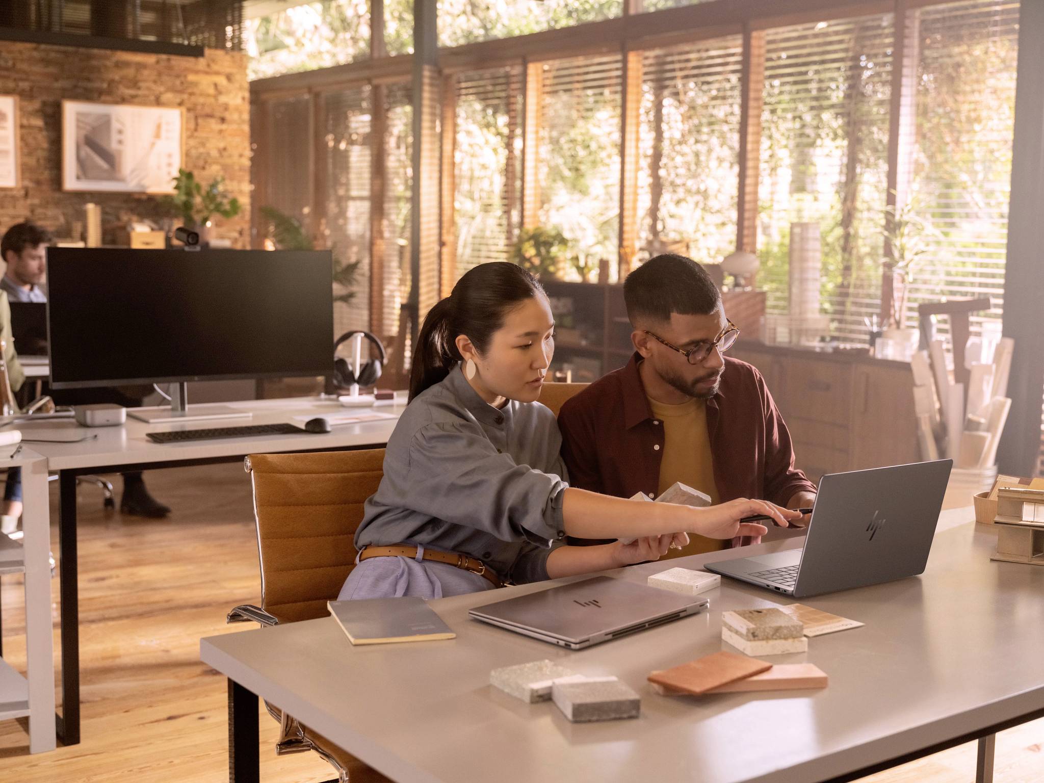 Deux personnes travaillent ensemble sur un ordinateur portable dans un bureau moderne avec des sols en bois et de grandes fenêtres. Des ordinateurs et des étagères de classement sont visibles en arrière-plan.
