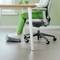 Une personne est assise sur une chaise de bureau à une table et utilise un entraîneur de pieds ergonomique. Un mur vert est visible en arrière-plan.
