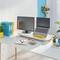 Bureau moderne avec un ordinateur portable et un écran, entouré de plantes. À côté se trouvent des dossiers colorés et du matériel de bureau.