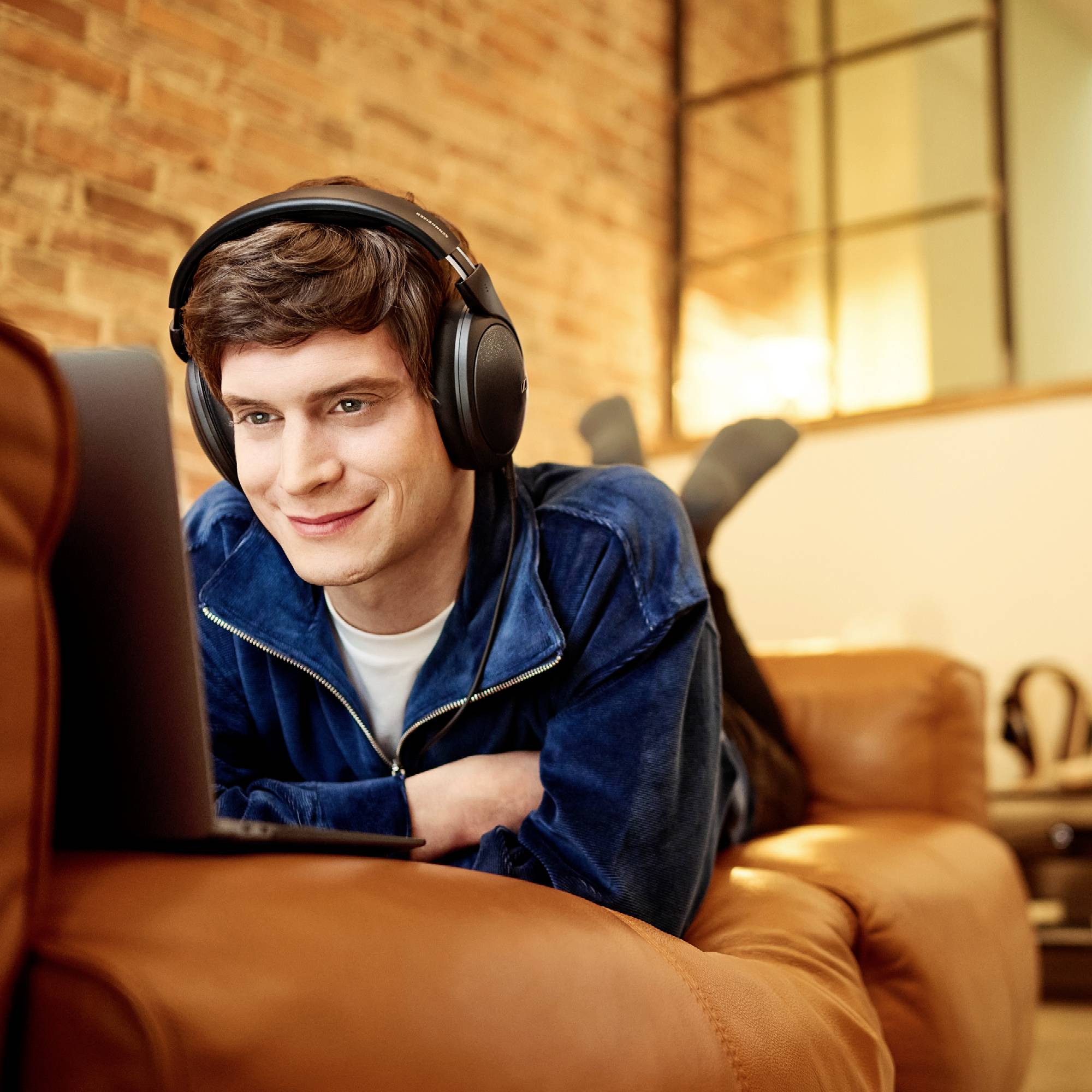 Un jeune homme est allongé sur un canapé, portant des écouteurs et regardant son ordinateur portable. Il a l'air détendu et concentré.