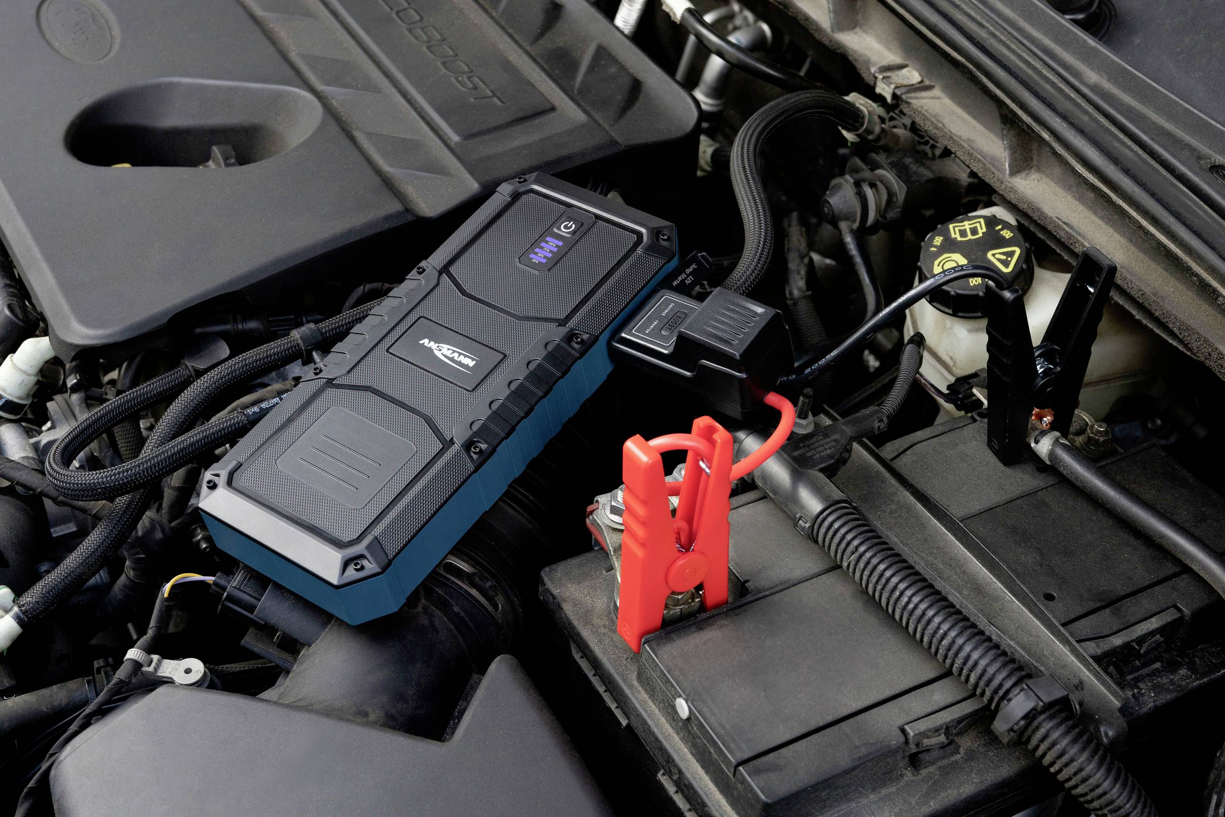Batterie de voiture dans un compartiment moteur avec un câble de démarrage rouge branché. Un appareil noir est posé à côté de la batterie.