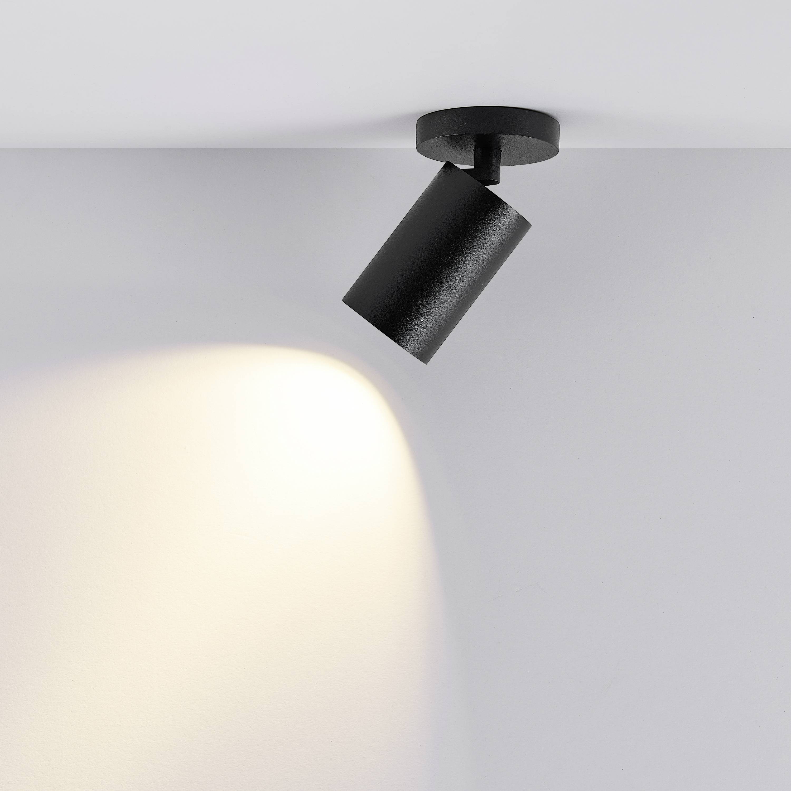 Projecteur de plafond noir éclairant une zone du mur avec une lumière blanc chaud. Design moderne et minimaliste.
