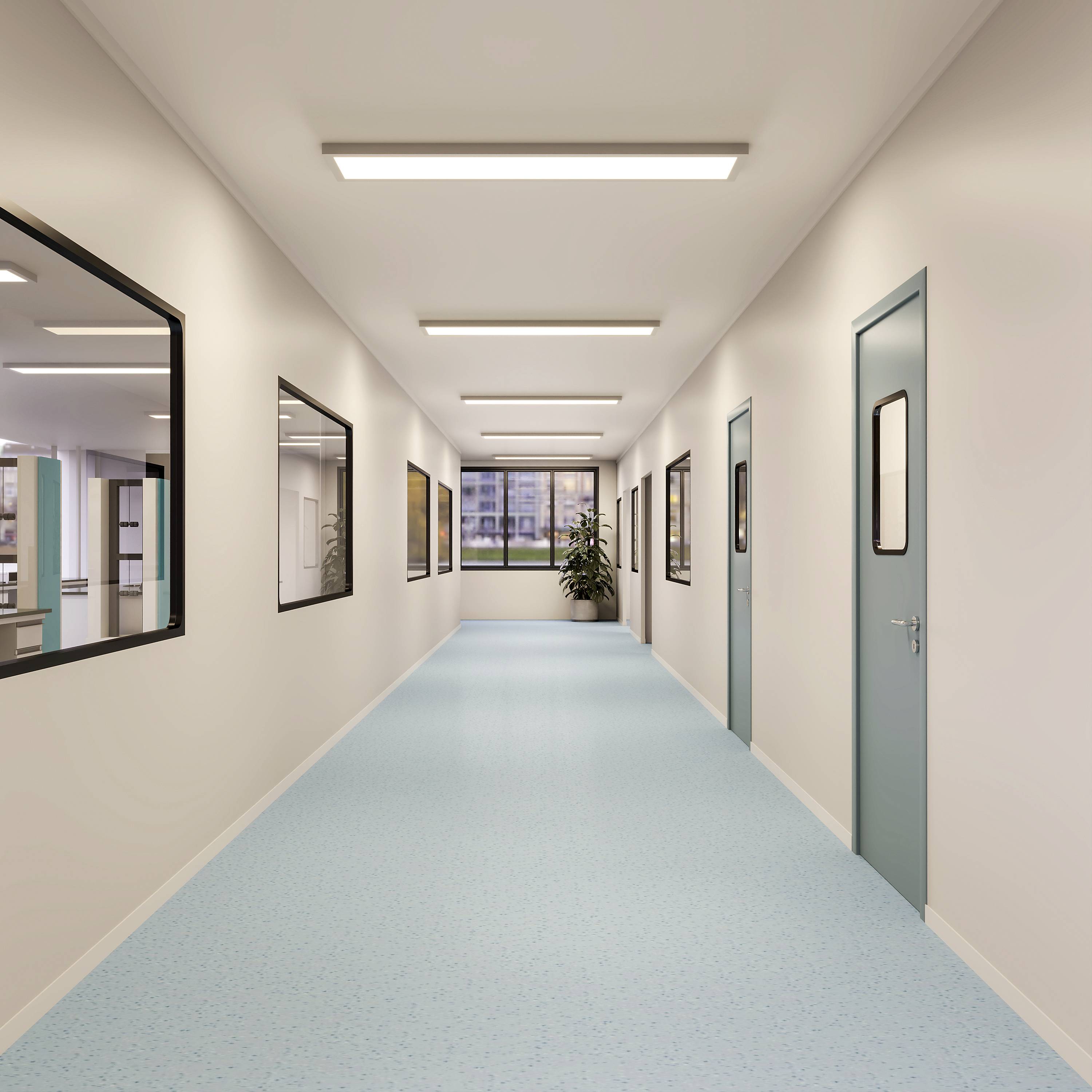 Un couloir d'hôpital vide avec un sol bleu clair et des portes des deux côtés ; de grandes fenêtres au bout laissent entrer la lumière du jour.