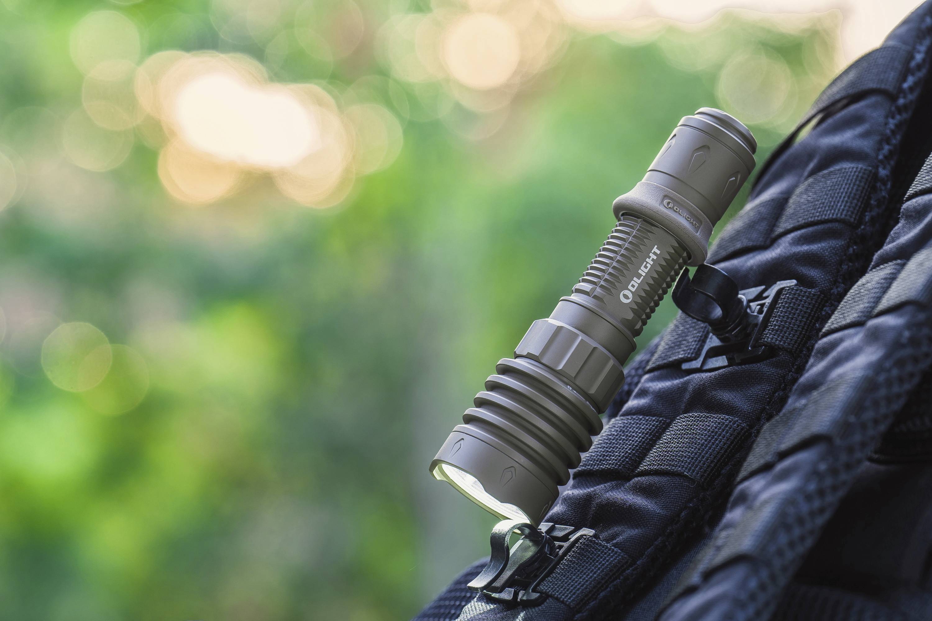 Une lampe de poche grise est fixée à un sac à dos noir. L'arrière-plan montre une nature verdoyante floue.