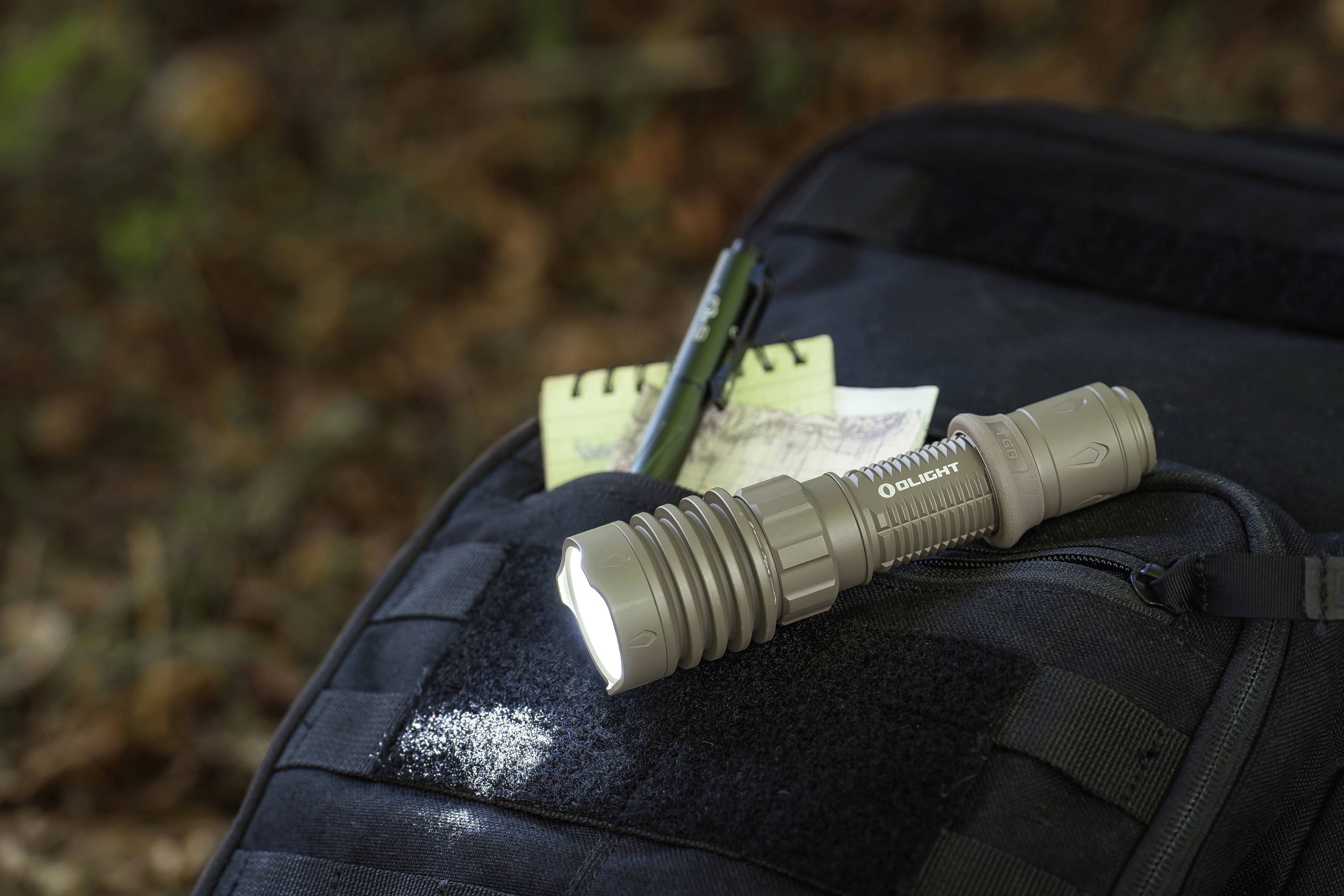 Une lampe de poche allumée est posée sur un sac à dos noir, à côté d'un carnet de notes et d'un stylo, en extérieur.