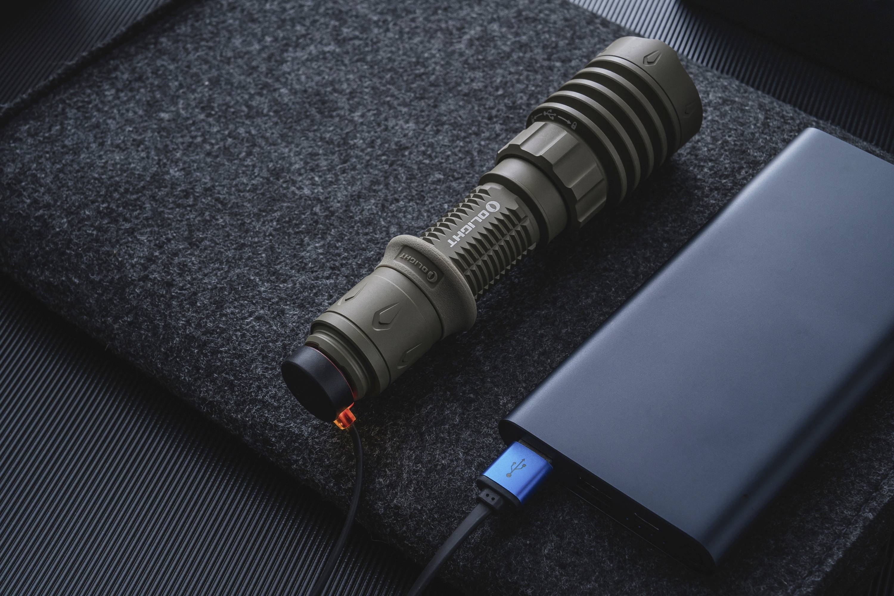 La lampe de poche se charge via un câble USB connecté à une batterie externe. La lampe de poche est posée sur une surface sombre en tissu.