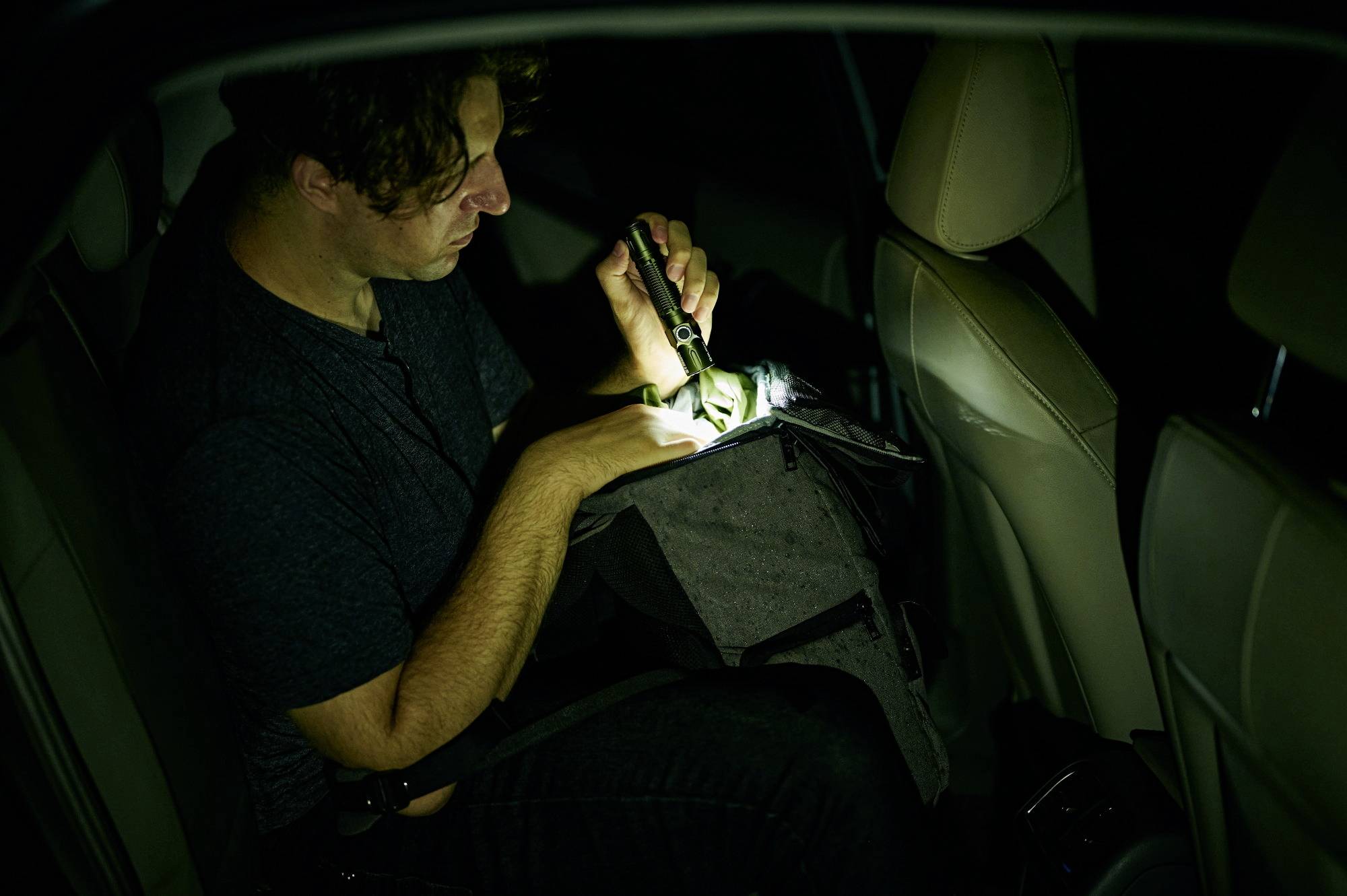 Un homme est assis dans sa voiture et braque une lampe de poche sur un sac. Il fait sombre, et il semble chercher quelque chose.