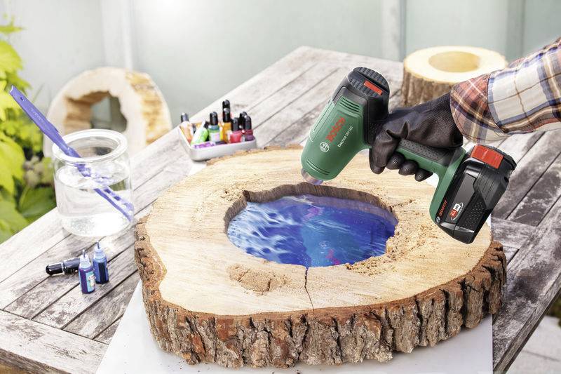 Un artisan utilise un pistolet à air chaud pour travailler la résine époxy dans un disque de bois. Sur la table sont disposés des résines et des couleurs.