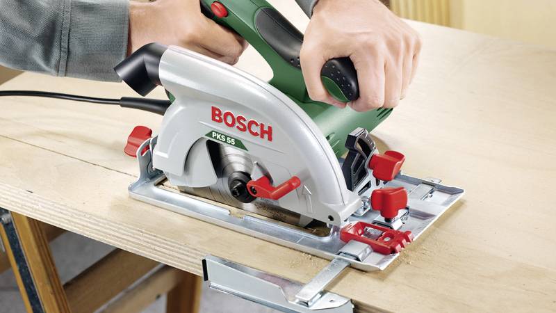 Une main tient une scie circulaire électrique verte de la marque 'Bosch' et coupe précisément à travers une planche en bois.