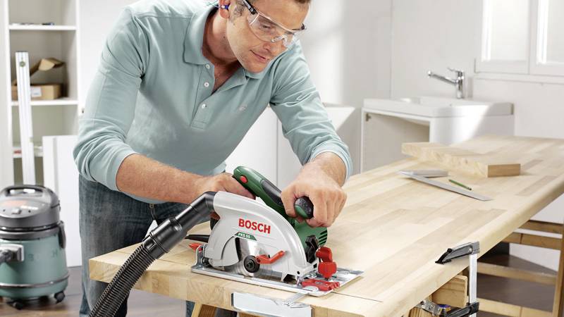 Un homme utilise une scie circulaire Bosch sur un établi, porte des lunettes de protection, dans un atelier avec un aspirateur en arrière-plan.