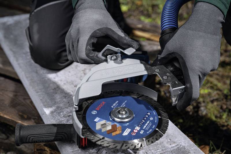 Une personne portant des gants gris fixe un capot de protection sur un disque de coupe Bosch bleu, posé sur un support métallique.