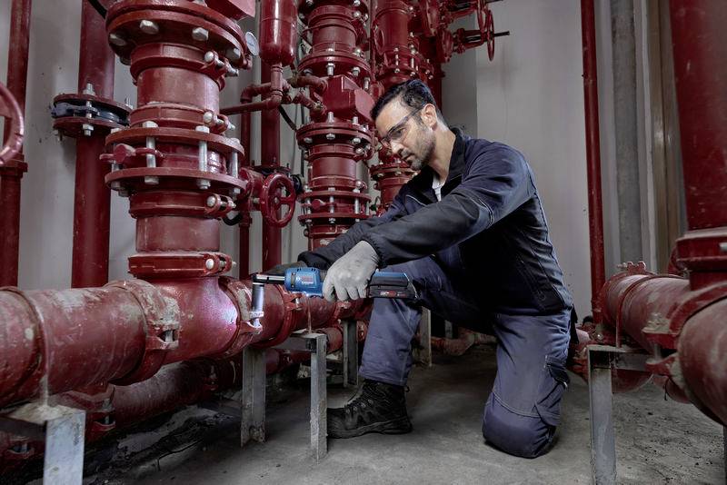 Un technicien répare un système de tuyauterie rouge dans un bâtiment industriel. Il porte des lunettes de protection et des gants de travail.