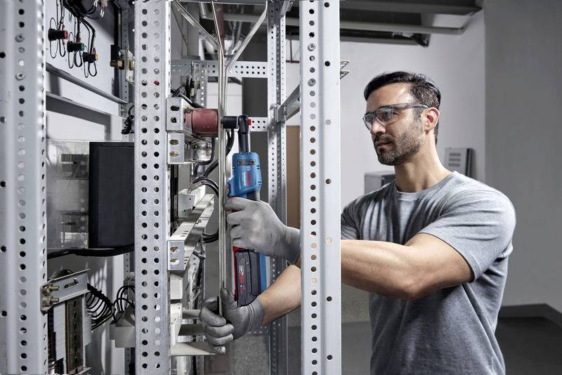 Un homme travaille dans un armoire électrique, porte des lunettes de protection et des gants, et utilise un outil électrique.