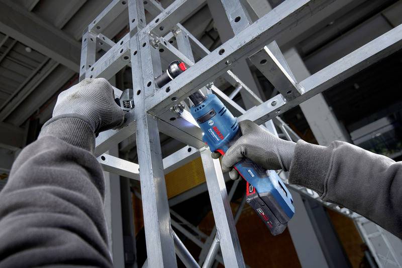 Une personne monte une structure métallique à l'aide d'un tournevis électrique bleu. Les mains portent des gants gris.