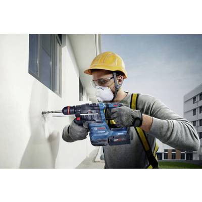 Un ouvrier portant un casque et un masque de protection fore un mur blanc avec une perceuse électrique sur un bâtiment moderne.