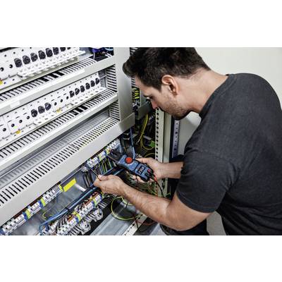 Un homme répare un armoire électrique. Il tient un appareil de mesure et vérifie les connexions de câbles.