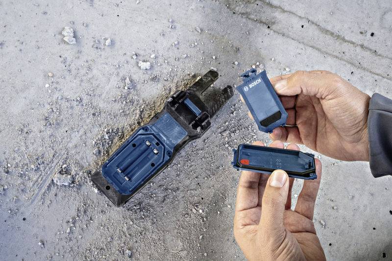 Deux mains tiennent un embout de vissage Bosch et une batterie. Un autre outil Bosch est posé sur un sol de type béton.