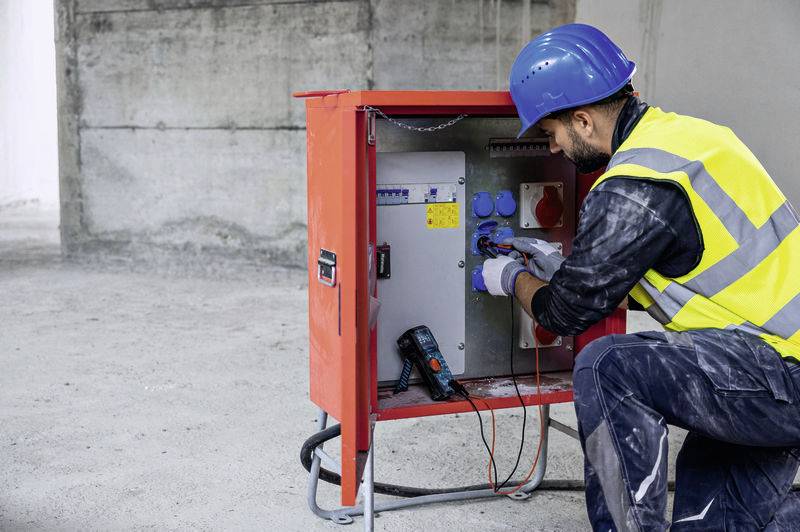 Un travailleur portant un casque de protection bleu et un gilet de sécurité vérifie un boîtier de tableau électrique sur un chantier de construction.