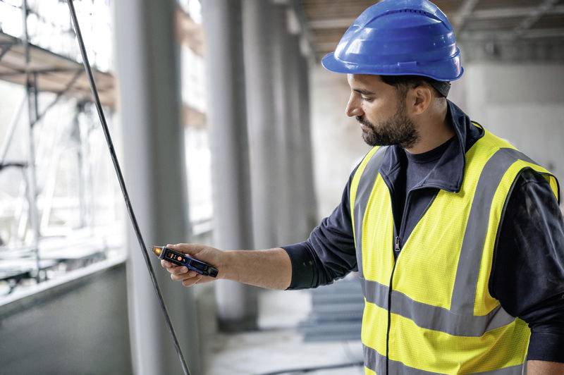 Un ouvrier portant un gilet de sécurité jaune et un casque de protection bleu vérifie un échafaudage à l'aide d'un appareil à l'intérieur d'un chantier de construction.