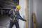 Un ouvrier portant un casque jaune travaille avec une scie électrique sur un pilier en béton sur un chantier. Un gilet de sécurité est visible.