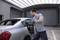 Un homme en tenue de travail scie la lunette arrière d'une voiture argentée avec une scie électrique dans un atelier ou un garage.