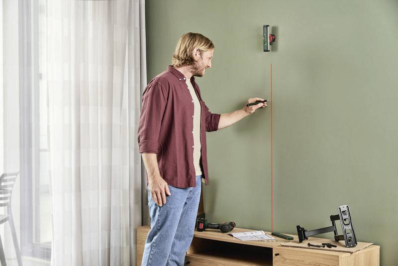 Une personne mesure la hauteur sur un mur à l'aide d'un outil laser. Des plans de construction et une perceuse sont posés sur une table.