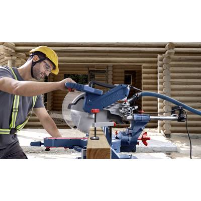 Un ouvrier portant un casque de chantier et des lunettes de protection utilise une scie circulaire sur table dans un chantier de construction en bois. Des poutres en bois sont visibles en arrière-plan.