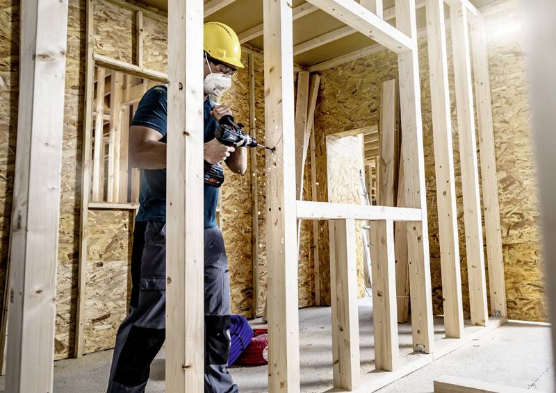 Un ouvrier porte un casque de chantier jaune et un masque de protection tout en travaillant avec une perceuse sur un chantier de construction de maison à ossature bois.