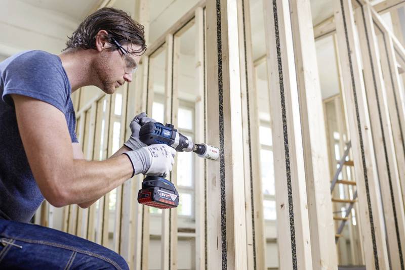 Un homme portant des lunettes de protection et des gants perce un trou dans un cadre en bois à l'aide d'une perceuse sans fil dans une pièce en cours de rénovation.