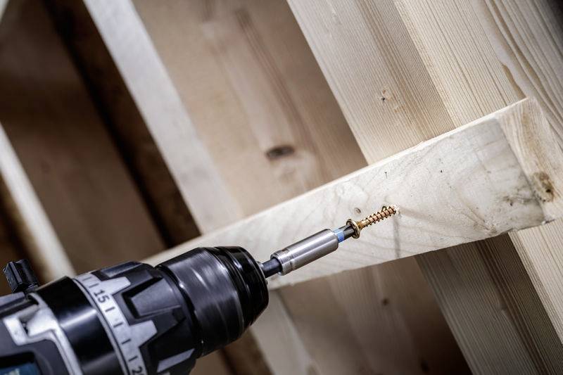 Une perceuse visse une vis dans une planche de bois. La structure du bois est visible, montrant un travail artisanal.