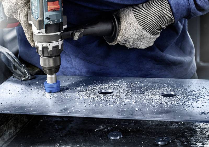 Une personne portant des gants de protection perce un trou dans une tôle métallique à l'aide d'une machine. Des copeaux de métal sont répartis autour du point de perçage.