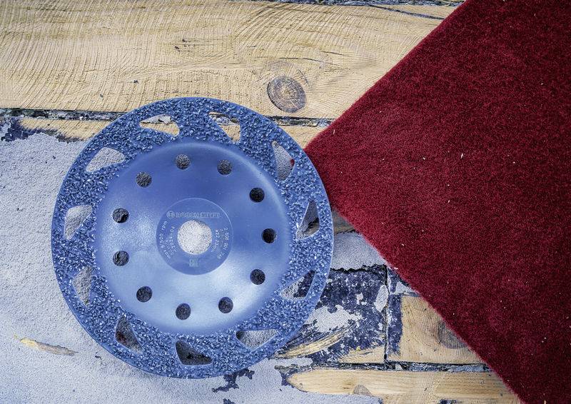 Un disque abrasif bleu est posé sur un sol en béton brut à côté d'un tapis rouge. Le disque abrasif est utilisé pour lisser des surfaces.