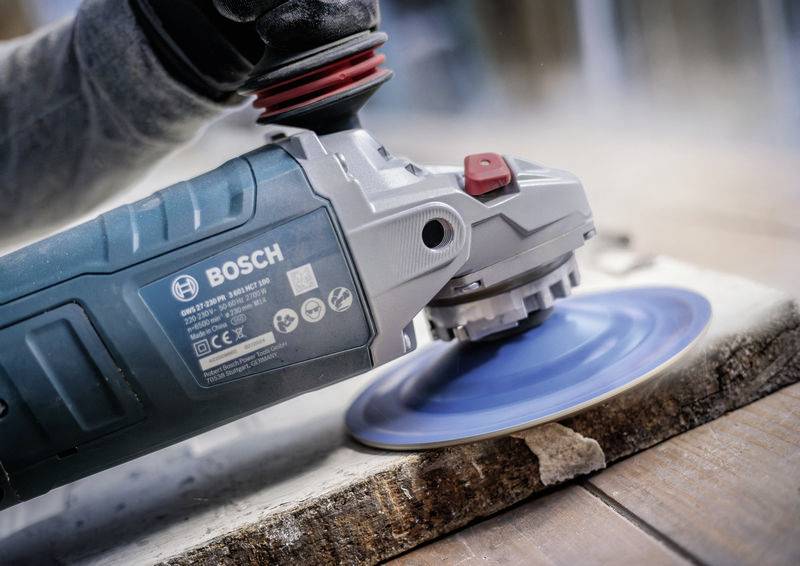 Une personne ponce une surface en bois avec une meuleuse d'angle de marque Bosch, utilisant un disque abrasif bleu.