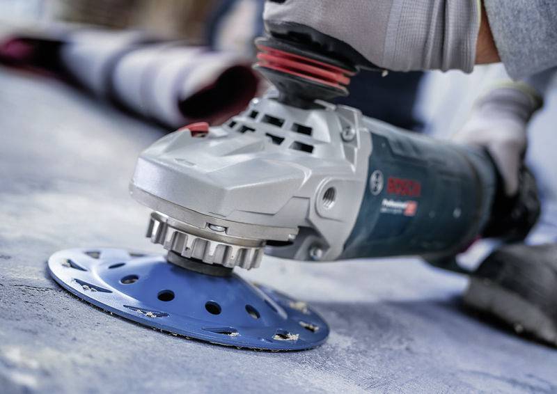 Une main tient une meuleuse électrique avec un disque de ponçage bleu. Elle ponce une surface en béton.