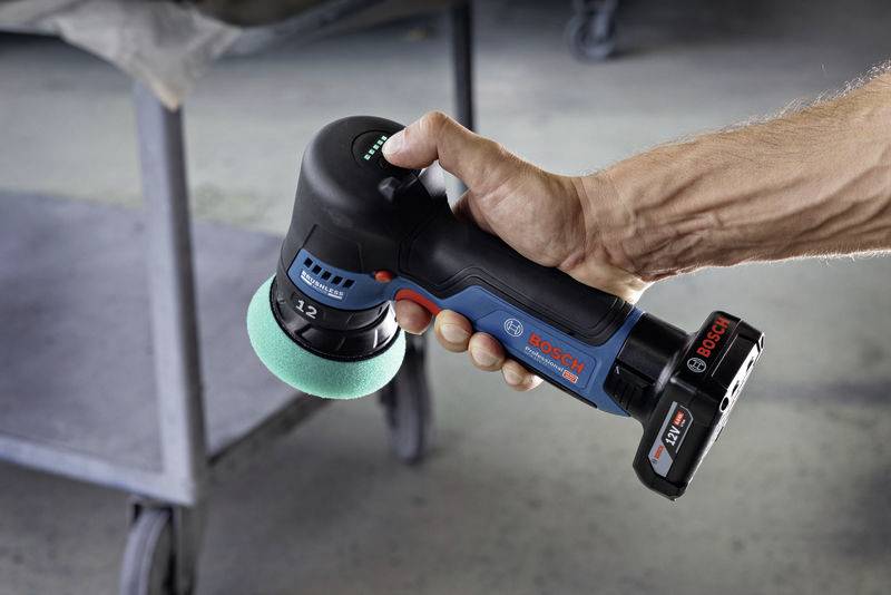 Un polisseur à batterie est tenu par une main. Il porte le logo de marque 'Bosch' et est utilisé dans un atelier.