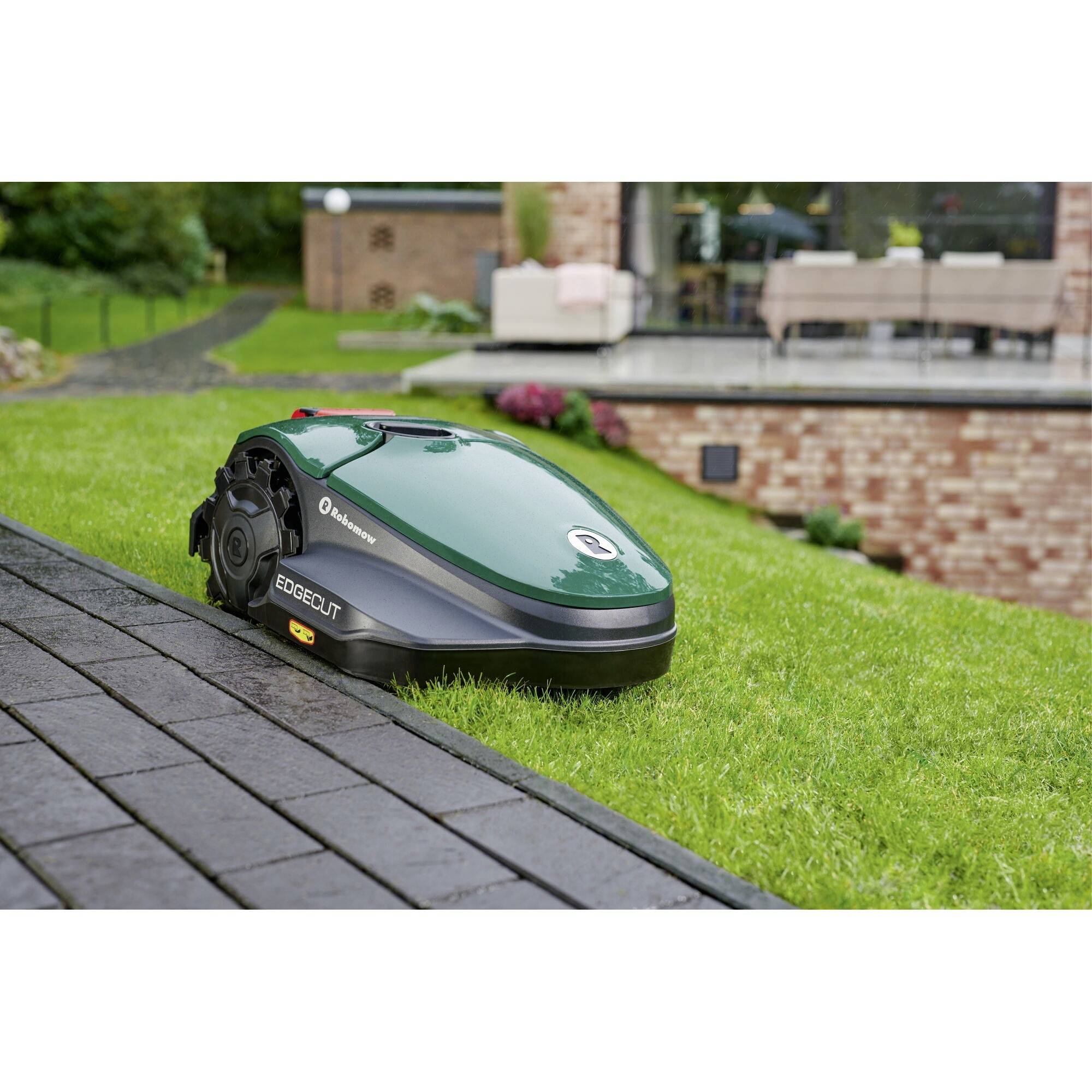 Un robot tondeuse tond un jardin, près d'une terrasse meublée. Scène lumineuse de jour, avec l'accent mis sur l'appareil autonome sur la pelouse.