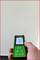 Une main tient un télémètre laser vert pointé vers un mur blanc. Un petit point rouge indique la position du laser.
