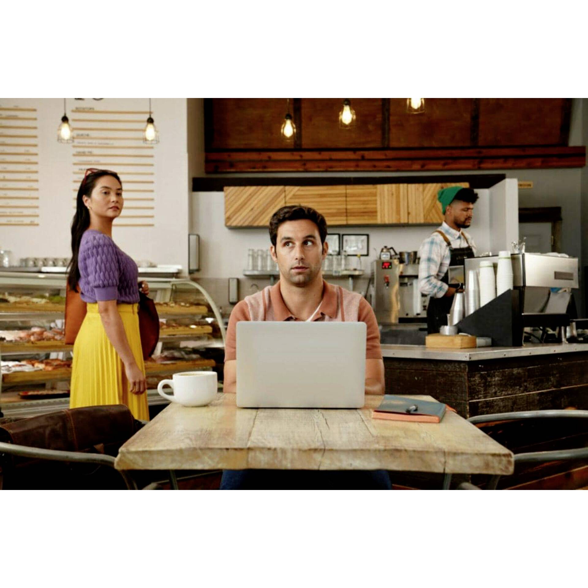 Un homme est assis à une table de café devant un ordinateur portable, avec une expression de surprise sur le visage. Une femme portant une jupe jaune le regarde.